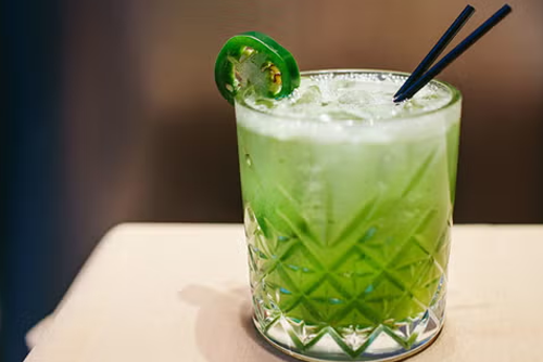 a green cocktail garnish in a glass with a jalapeño slice on the rim